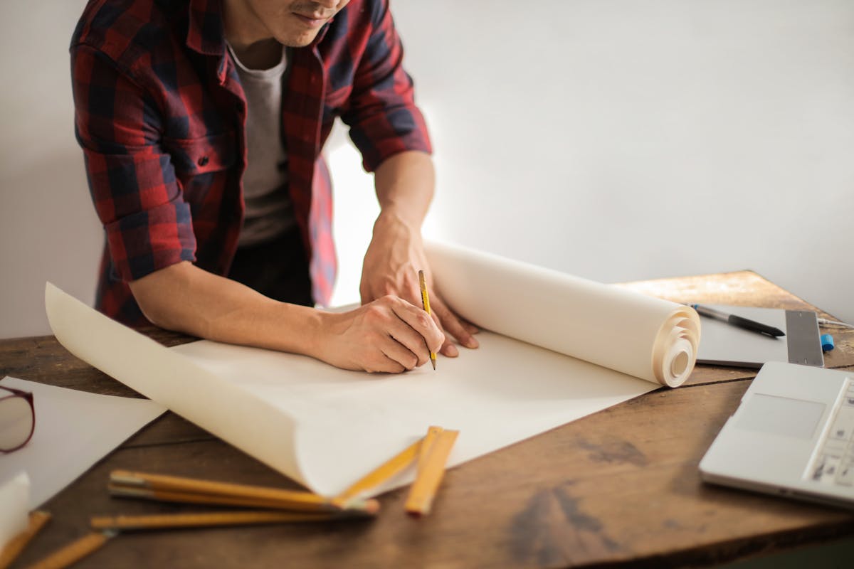 Person sketching architectural designs on a large paper roll at a wooden desk with rulers and a laptop
