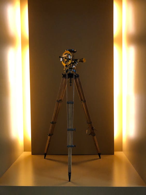 Antique brass theodolite survey instrument displayed on a wooden tripod with dramatic lighting