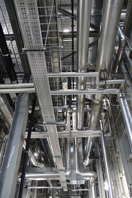 Dense network of steel pipes, conduit, and cable trays running through an industrial building ceiling