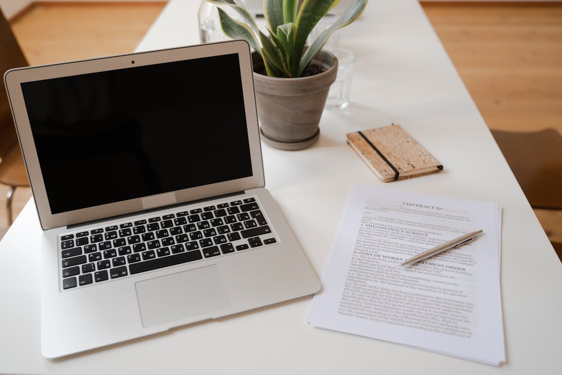 Laptop computer on a desk next to contract documents with a pen and notebook