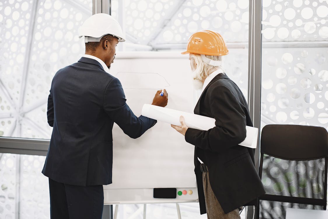 Two professionals in hard hats reviewing architectural plans on a whiteboard in an office