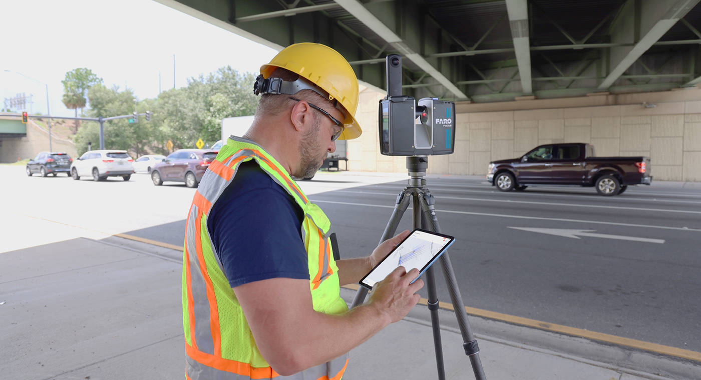 Technician in safety vest and hard hat operating a FARO Focus laser scanner with tablet controller at an infrastructure site