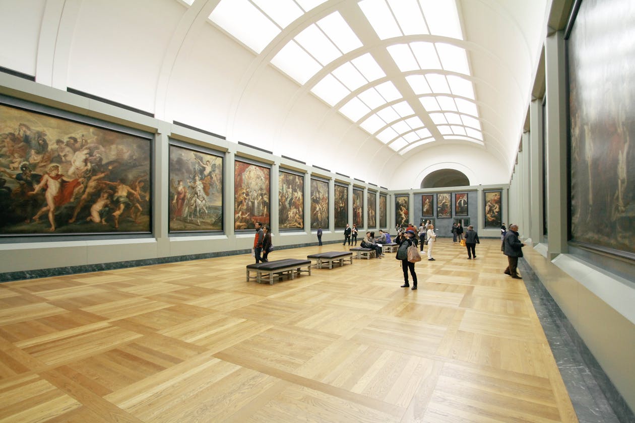 Visitors walking through a grand museum gallery with large classical paintings and natural skylight illumination