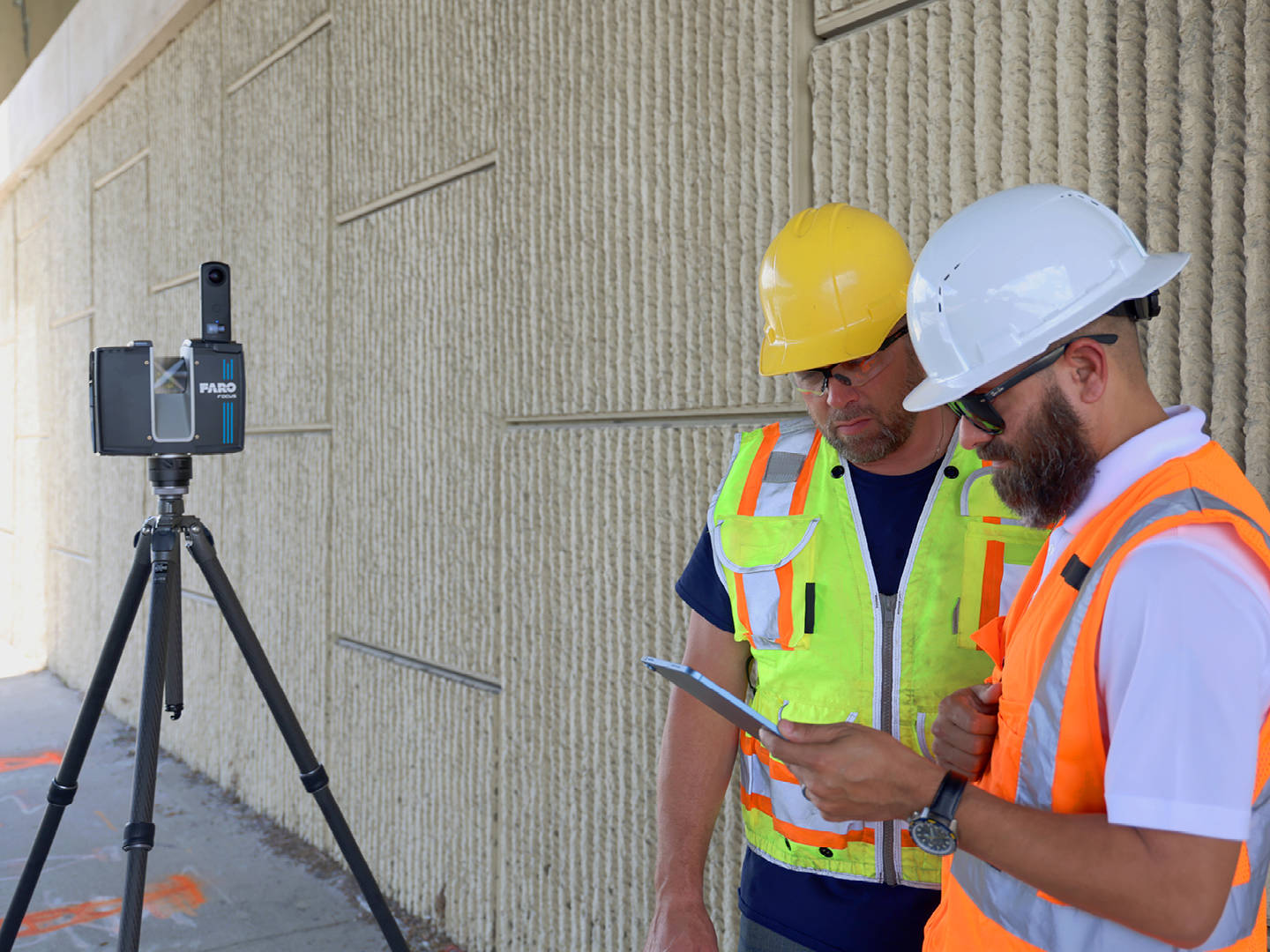 Two construction professionals in safety vests and hard hats reviewing scan data on a tablet next to a FARO Focus laser scanner on a tripod at a construction site