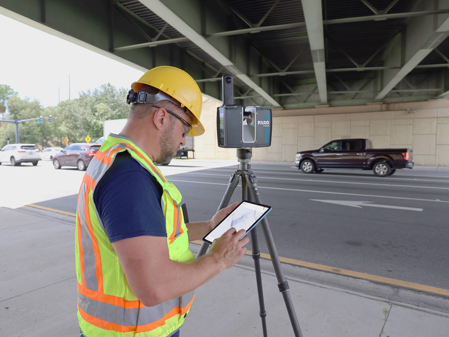 Surveyor in safety vest and hard hat operating a FARO Focus laser scanner on a tripod under a bridge, reviewing scan data on a tablet