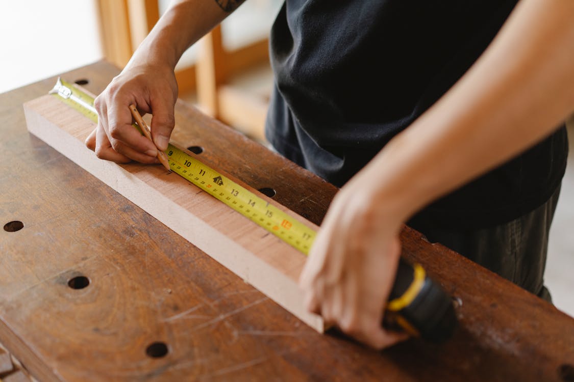 Hands measuring a wooden plank with a yellow tape measure and pencil on a workbench