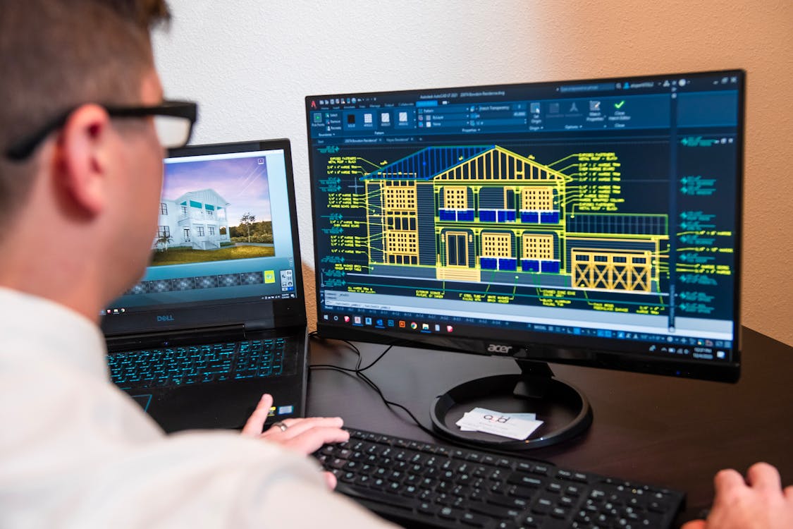 Architect working at dual monitors with AutoCAD showing building structural wireframe alongside a 3D rendering