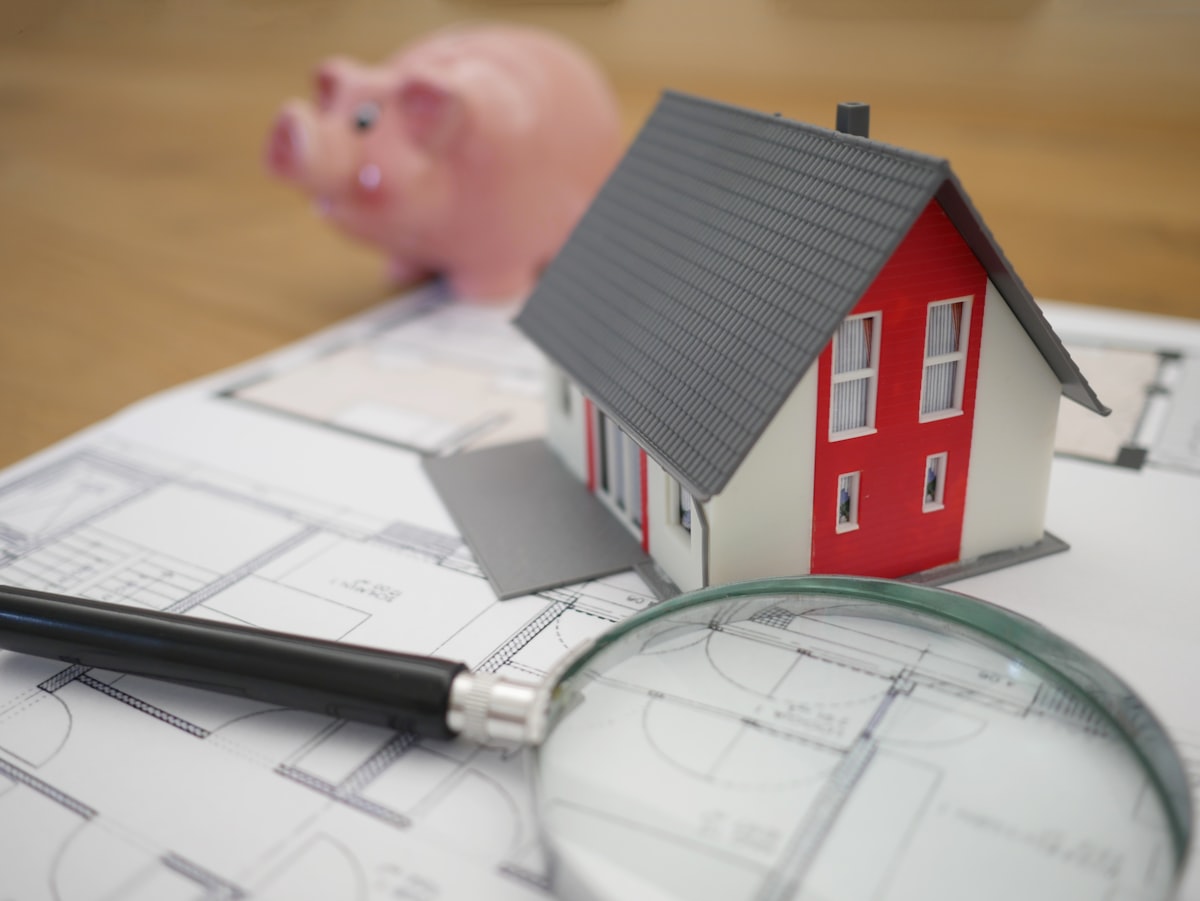 Miniature house model on architectural floor plans with magnifying glass and piggy bank, representing real estate investment and property technology