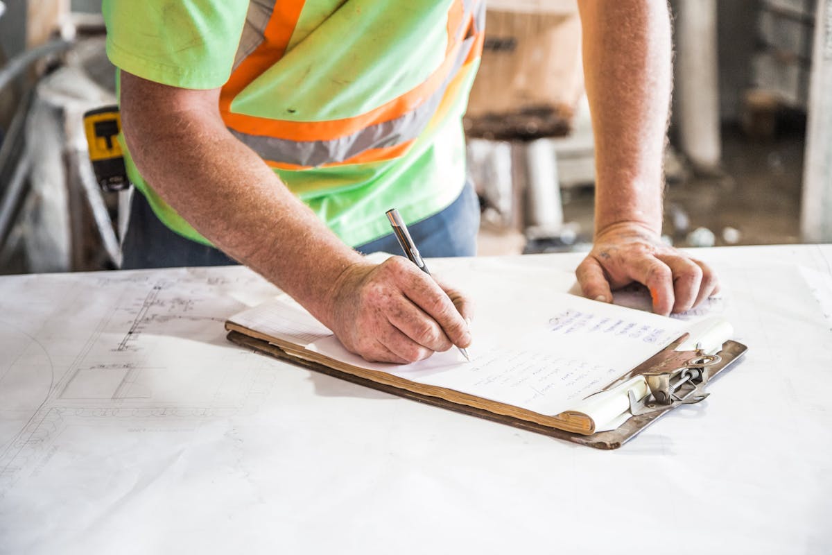 Construction worker in safety vest writing notes on a clipboard with blueprints spread on a work surface