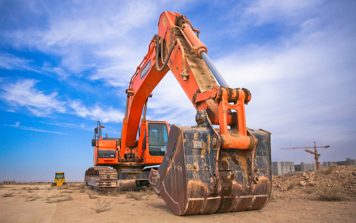 Orange excavator at a construction site with cranes visible in the background under blue sky